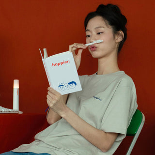 White diary notebook held in hand, cover shows "happier." and face illustration