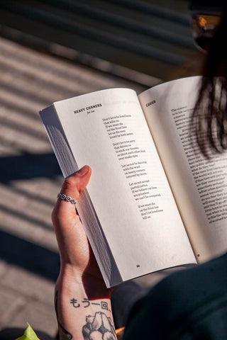 A person with black ink tattoos on their wrist and a silver ring with daisy chain design holds a poetry book in the afternoon sun. We see the page they look at.