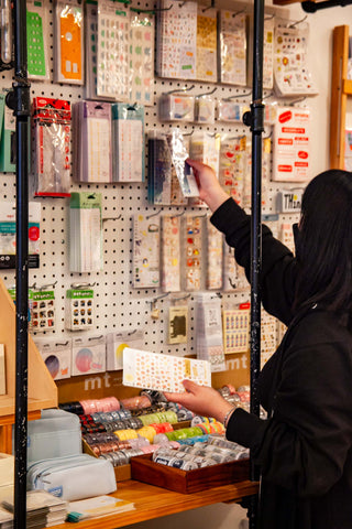 A photo of M restocking a wall full of stickers on the OMOI sales floor. Their left hand is full of stickers.