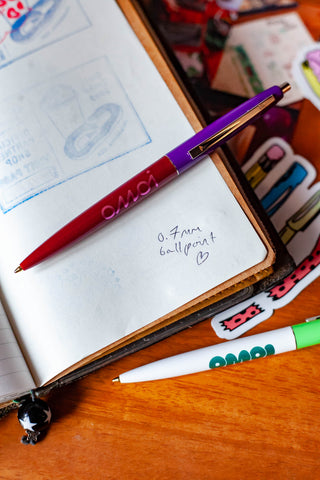 OMOI Shop Pen with dark berry body and purple cap, lying on notebook