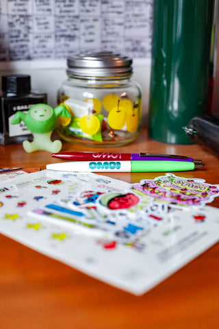 OMOI Shop Pen set: dark berry and key lime pens on a desk beside stickers