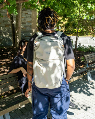 A person standing outdoors on a paved walkway, viewed from behind, wearing a light beige backpack. Another person sits on a bench in the background under dappled sunlight.