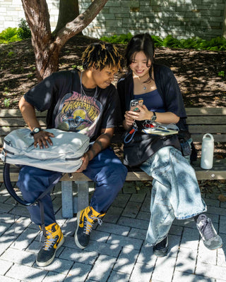 Two people sitting on a park bench looking at a phone, with a light gray backpack resting on the bench in the foreground.