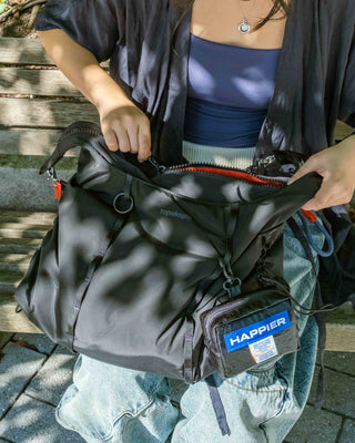A person outdoors holding open a black bag, showing its interior and a small attached pouch labeled HAPPIER. The scene includes a bench and dappled sunlight