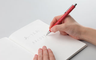 STÁLOGY 015 red ballpoint pen writing in an open notebook