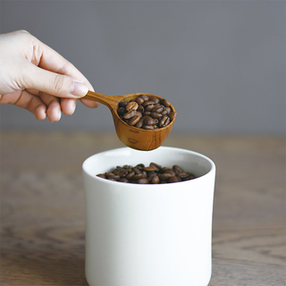 Wooden coffee measuring spoon holding coffee beans above a container
