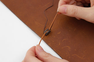 Hands repairing a brown notebook cover seam with a thin cord and needle tool