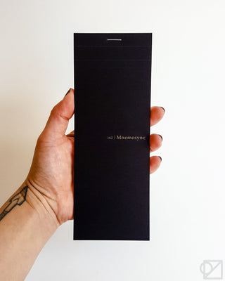 Black grid notepad held in hand, gold "Mnemosyne" logo on cover