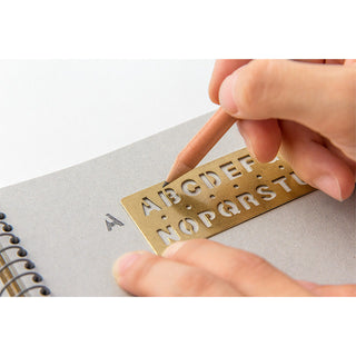 Brass alphabet template bookmark held to paper for tracing A–Z letters