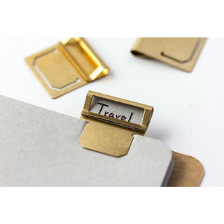Brass index clip on gray surface, stamped 'Travel' tag with hinged metal top