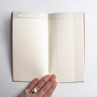 Open Traveler's notebook showing dated grid pages with fingers resting on paper
