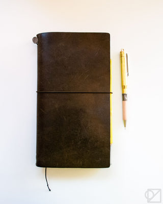 Brass ballpoint pen beside a black leather notebook with notebook strings