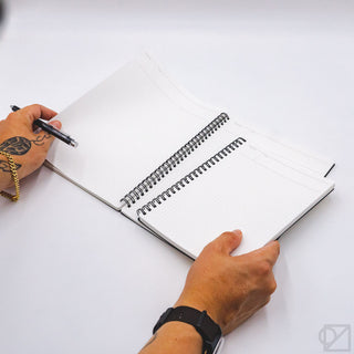 Opened white dot-grid spiral notebook held with hands and black pen