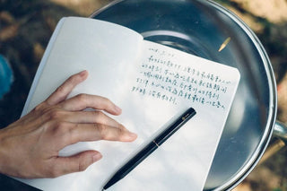 Hands writing with a black ballpoint pen on paper atop a metal tray